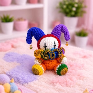 Könnte beinhalten: Eine gehäkelte Narrenpuppe mit weißem Gesicht und schwarzen Kreuzaugen, einer blau-violetten zweizipfligen Mütze mit goldenen Sternanhängern und einem regenbogenfarbenen Rüschenkragen. Der Körper ist orange mit gelb-grünen strukturierten Füßen.