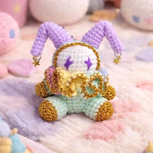 May include: A handmade crochet doll in pastel colors. The doll has a white face with purple accents, lavender ears, and gold trim. It is adorned with a gold star charm and is sitting on a soft, multi-colored surface. Other crochet items are in the background.