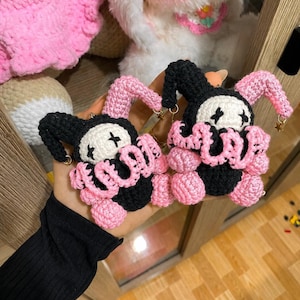 May include: Two handmade crochet plush keychains featuring a jester-like character with a white face, black X-shaped eyes, and a ruffled collar. One figure wears a black hat with pink accents, while the other wears a pink hat with black accents.