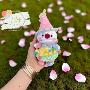 May include: A handmade crochet clown doll with a pink hat, white face, and colorful outfit. The doll has a red nose, black eyes, and a yellow, blue, and green outfit. The doll is held in a hand, with a background of green moss and pink flower petals. The logo "Lily Craft House" is in the top left corner.