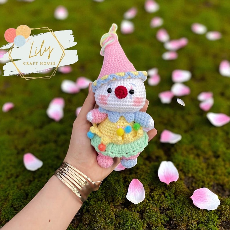 May include: A handmade crochet clown doll with a pink hat, white face, and colorful outfit. The doll has a red nose, black eyes, and a yellow, blue, and green outfit. The doll is held in a hand, with a background of green moss and pink flower petals. The logo "Lily Craft House" is in the top left corner.