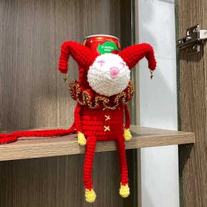 Puede incluir: Muñeca de bufón roja y blanca hecha a mano con ganchillo, con cola roja, pies amarillos y un collar dorado. El bufón tiene una cara blanca con una nariz rosa y lleva un sombrero rojo con detalles de estrellas doradas. Una lata se encuentra encima de la muñeca.