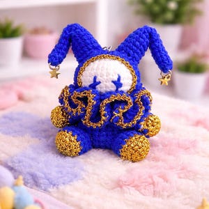 May include: A handmade blue and gold crochet doll with a whimsical design. The doll features a white face with blue cross-shaped eyes, a ruffled collar, and star-shaped charms. It is set against a soft, pastel-colored background.