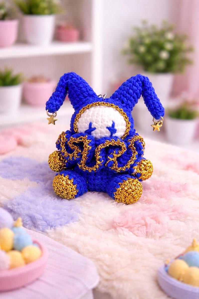 May include: A handmade blue and gold crochet doll with a whimsical design. The doll features a white face with blue cross-shaped eyes, a ruffled collar, and star-shaped charms. It is set against a soft, pastel-colored background.