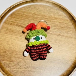 May include: A handmade crochet clown keychain. The clown has a red and black checkered outfit, a green collar, and a colorful hat with red and orange accents. The keychain is on a wooden tray.