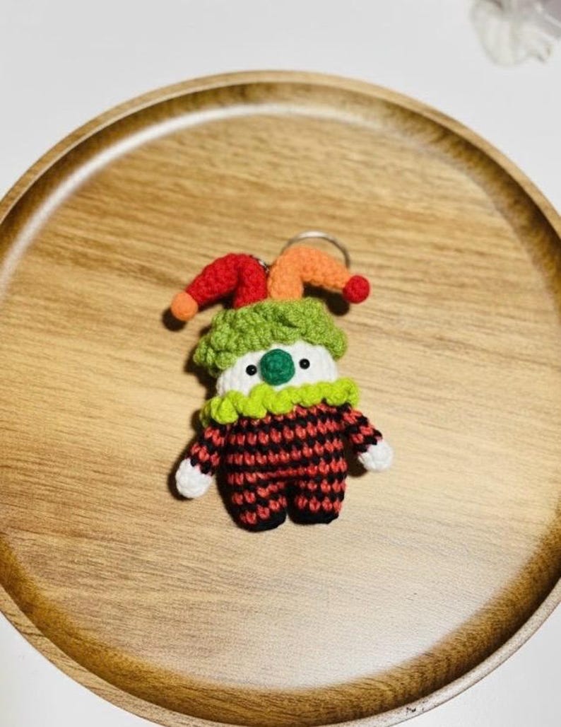 May include: A handmade crochet clown keychain. The clown has a red and black checkered outfit, a green collar, and a colorful hat with red and orange accents. The keychain is on a wooden tray.