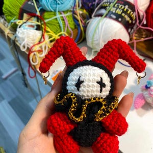 May include: A handmade crochet jester doll with a red and black outfit, white face, and gold accents. The doll has red horns and is adorned with small gold-colored hoop earrings. The background includes colorful yarn skeins.