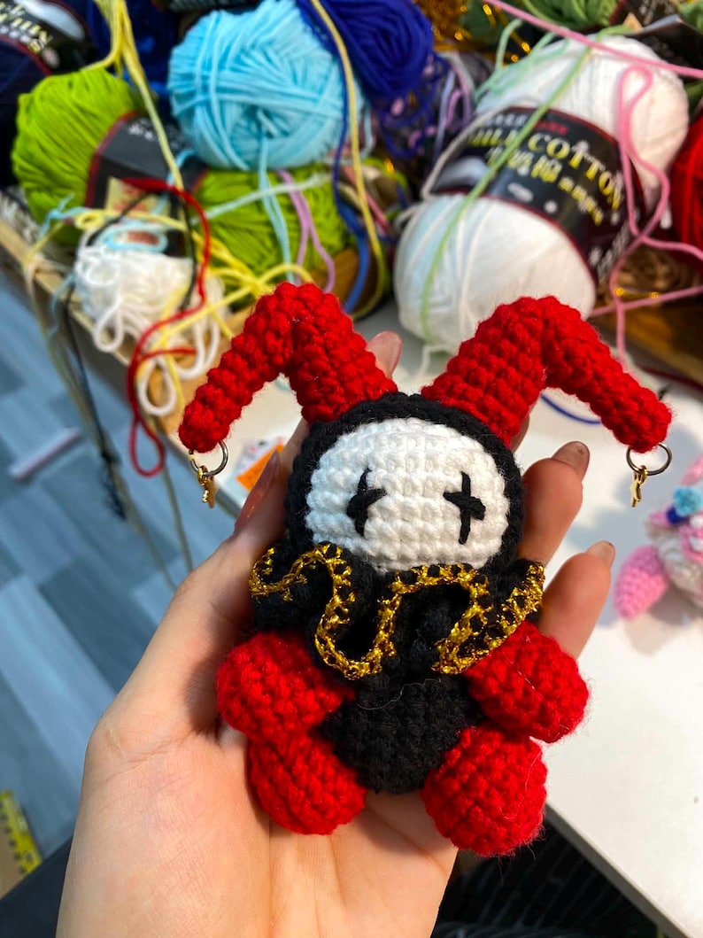 May include: A handmade crochet jester doll with a red and black outfit, white face, and gold accents. The doll has red horns and is adorned with small gold-colored hoop earrings. The background includes colorful yarn skeins.