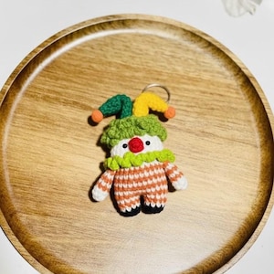 May include: A handmade crochet clown keychain. The clown has a white face with a red nose, a green and orange striped outfit, and a green and yellow jester hat. The keychain is on a wooden tray.