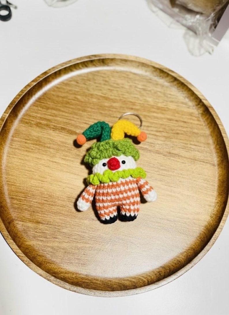 May include: A handmade crochet clown keychain. The clown has a white face with a red nose, a green and orange striped outfit, and a green and yellow jester hat. The keychain is on a wooden tray.