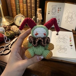 May include: A handmade crochet doll in the shape of a jester. The doll has a green body, a white face with red accents, and a brown ruffled collar. The jester's hat is red with star-shaped embellishments.