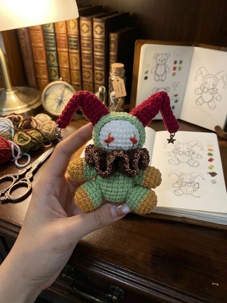 May include: A handmade crochet doll in the shape of a jester. The doll has a green body, a white face with red accents, and a brown ruffled collar. The jester's hat is red with star-shaped embellishments.
