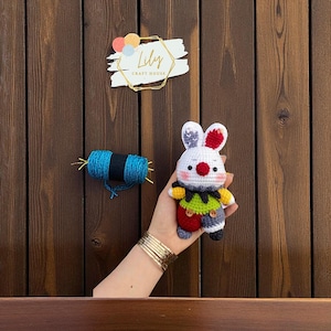 May include: A handmade crochet bunny doll with a white body, red and gray ears, and a colorful outfit. The doll is held in a hand, with a spool of blue yarn and a "Lily Craft House" sign in the background.