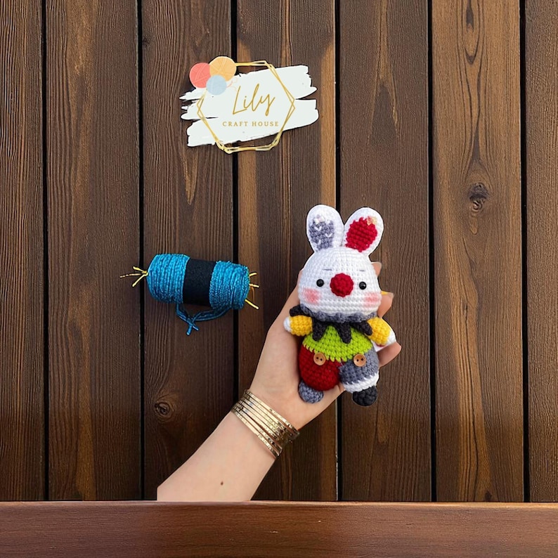 May include: A handmade crochet bunny doll with a white body, red and gray ears, and a colorful outfit. The doll is held in a hand, with a spool of blue yarn and a "Lily Craft House" sign in the background.