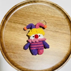 May include: A handmade crochet clown keychain. The clown has a yellow, purple, and pink jester hat with orange pom-poms. The clown's body is purple and pink striped, with purple arms and legs. It has a red nose and white face.