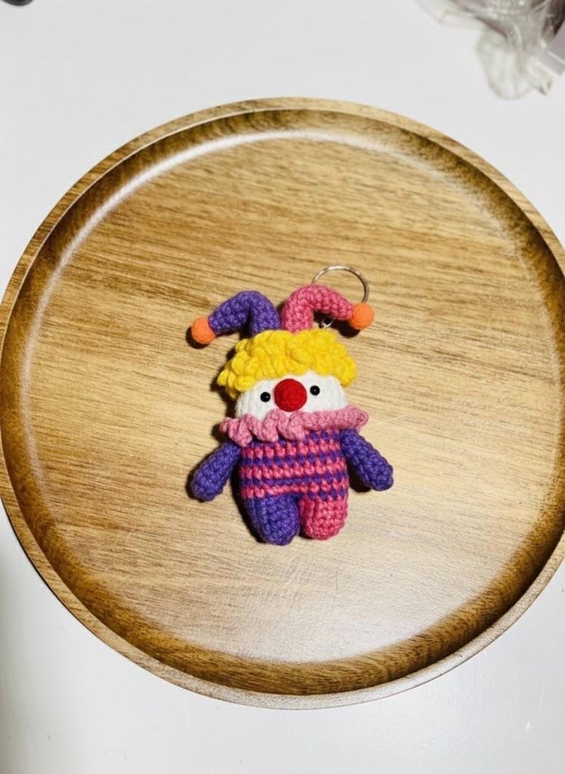 May include: A handmade crochet clown keychain. The clown has a yellow, purple, and pink jester hat with orange pom-poms. The clown's body is purple and pink striped, with purple arms and legs. It has a red nose and white face.