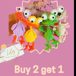 Puede incluir: Una colección de llaveros de crochet hechos a mano con forma de animales caprichosos, incluyendo un pájaro morado, un pato amarillo, una rana verde y una criatura beige. Los llaveros tienen ojos grandes y están unidos a llaveros plateados. Se muestra el texto "Buy 2 get 1".