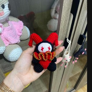May include: A handmade crochet jester doll with a red and black color scheme. The doll has a white face with red accents, a gold collar, and red jester hat. Other crochet items are visible in the background.
