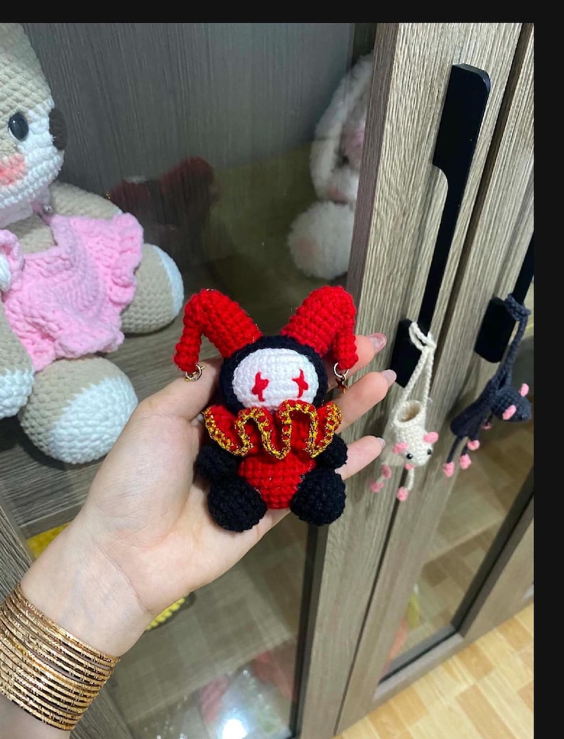 May include: A handmade crochet jester doll with a red and black color scheme. The doll has a white face with red accents, a gold collar, and red jester hat. Other crochet items are visible in the background.
