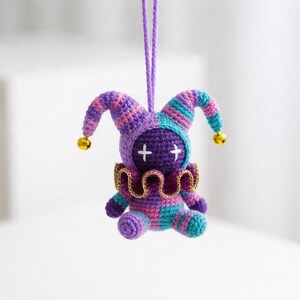 May include: A handmade crochet jester doll in purple, teal, and pink. The doll has a jester hat with bells, a gold collar, and cross-shaped eyes. It is suspended by a purple string.