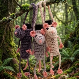 Könnte beinhalten: Drei handgefertigte, gehäkelte Mäusebeutel in Lila-, Grau- und Rosatönen, die an einem Ast hängen. Jede Tasche hat rosa Ohren, eine rosa Nase und herabhängende Beine. Die Beutel hängen vor einem verschwommenen Waldbild.
