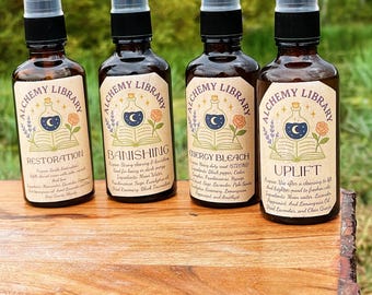 Op de afbeelding: Vier bruine glazen spuitflessen met zwarte sproeikoppen worden op een houten oppervlak weergegeven. Elke fles heeft een crèmekleurig etiket met de woorden "Alchemy Library" en een unieke naam: "Restoration", "Banishing", "Energy Bleach" en "Uplift".