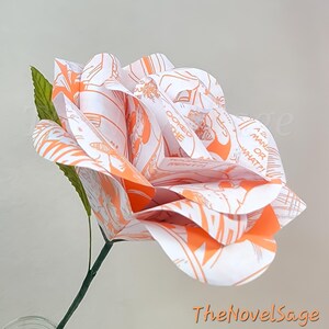 May include: A paper rose with white petals featuring orange comic book illustrations. The flower has a green stem and leaves. The text "The Novel Sage" is in the bottom right corner.