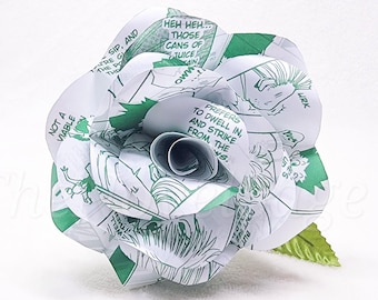 May include: A handmade paper rose crafted from comic book pages. The white petals feature green illustrations and text, with a green leaf and stem. The text includes phrases like "HEH HEH... THOSE CANS OF JUICE" and "PREFERS TO DWELL IN".