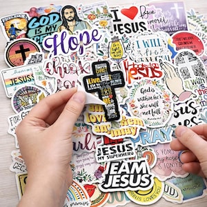 May include: A selection of vibrant stickers with religious motifs. The stickers display phrases such as "Jesus," "Hope," and "God is my," alongside images of crosses, Jesus, and inspirational quotes. A hand is peeling a sticker.