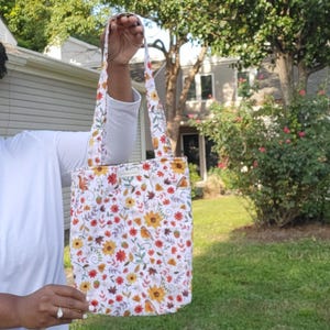 Könnte beinhalten: Eine weiße Tragetasche mit einem farbenfrohen Blumenmuster mit Sonnenblumen, roten Blumen und Herbstblättern. Die Tasche hat passende Griffe und ein kleines Etikett. Die Tasche wird vor einem Haus und grünem Laub gehalten.