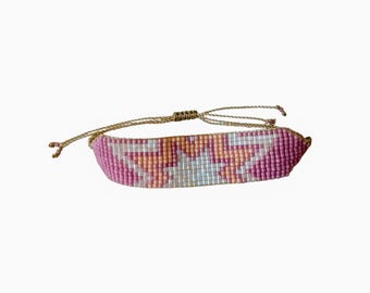 May include: A beaded bracelet with a geometric pattern in shades of pink, peach, and white. The bracelet is adjustable with a woven cord and a sliding knot closure. The bracelet is a fashion accessory.