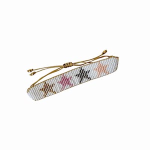 Op de afbeelding: Witte armband met gouden, roze en zwarte sterpatronen. De armband heeft een goudkleurige verstelbare koordsluiting. De sterren zijn gelijkmatig verdeeld over de witte kralenband. Een stijlvol accessoire.
