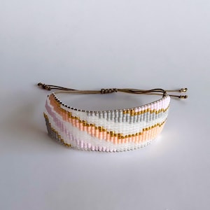 Könnte beinhalten: Gewebtes Armband mit einem sich wiederholenden Muster aus weißen, grauen, rosa und goldenen Perlen. Das Armband hat einen Schiebeknotenverschluss mit braunen Kordeln und goldfarbenen Endperlen. Modeschmuck.