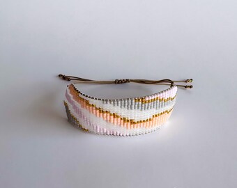 May include: Woven bracelet with a repeating pattern of white, gray, pink, and gold beads. The bracelet has a sliding knot closure with brown cords and gold-colored end beads. The bracelet is a fashion accessory.