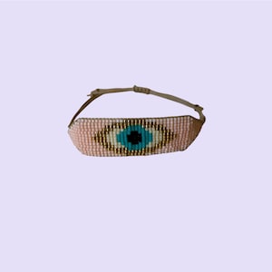 May include: A beaded bracelet featuring an eye design. The bracelet has a light pink background with a blue and black eye design, outlined in gold beads. The adjustable band is a light brown color.