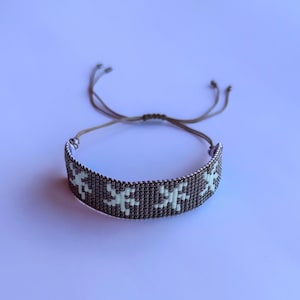 May include: A beaded bracelet with a repeating pattern of white shapes on a dark gray background. The bracelet is adjustable with a tan cord closure. The bracelet is shown on a light purple background.