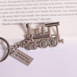 Può includere: Un portachiavi in metallo color argento con un ciondolo a forma di locomotiva a vapore e una targhetta rettangolare incisa con il testo My first hero, my forever dad. Il portachiavi include un anello circolare collegato da una catenella.