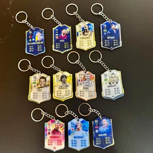 May include: A collection of soccer player keychains featuring various players like Cristiano Ronaldo, Messi, and Neymar. Each keychain has a player's image, name, and stats. The keychains are attached to silver key rings.