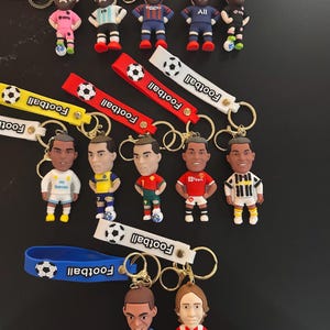 Puede incluir: Colección de llaveros con temática de fútbol, con figuras de jugadores en miniatura. Cada llavero tiene una correa colorida con la palabra "Football" y un diseño de balón de fútbol. Las figuras visten diferentes uniformes de equipo.