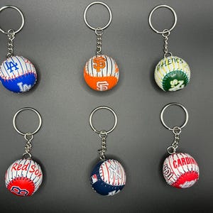 Puede incluir: Seis llaveros con temática de béisbol con aros plateados. Cada llavero presenta una minipelota de béisbol con logotipos de equipos, incluidos los Dodgers, Giants, Athletics, Red Sox, Yankees y Cardinals. Las pelotas son blancas con costuras rojas.