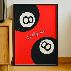 May include: A framed art print featuring two black and white 8-ball designs against a red background. The words "Lucky me" are written in white script. The artwork is in a black frame.