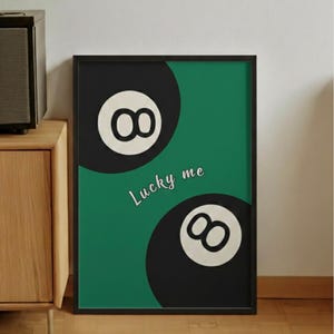 May include: A framed art print featuring two black eight-ball designs with white numerals against a green background. The words "Lucky me" are written in a cursive font. The artwork is in a black frame.