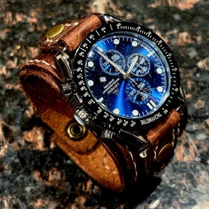 May include: A brown leather cuff watch with a blue face and black bezel. The watch features sub-dials and a tachymeter scale. The cuff is secured with brass rivets and a buckle.