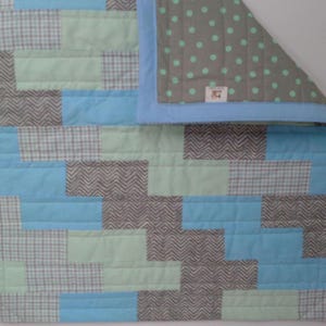 May include: A patchwork quilt featuring a mix of blue, gray, and mint green fabrics. The quilt's design includes rectangular blocks and a gray polka dot pattern on the reverse side. The edges are trimmed with a light blue border.