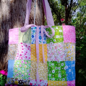 May include: A quilted tote bag featuring a patchwork design of various floral and gingham fabric squares in shades of pink, yellow, green, and blue. The bag includes two light pink shoulder straps and a matching light pink fabric tie closure.