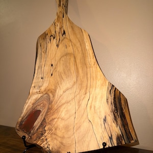 Puede incluir: Tabla de cortar de madera hecha a mano con una forma única y patrones de vetas naturales. La tabla presenta un asa y una incrustación de tono rojo. Se exhibe sobre un soporte de metal negro, mostrando su diseño artístico y funcionalidad.