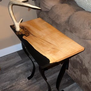 May include: A handcrafted side table with a light wood top and black base. The table features a natural wood grain and a decorative antler. The table measures approximately 30 inches long, 15 inches wide, and 24 inches high.