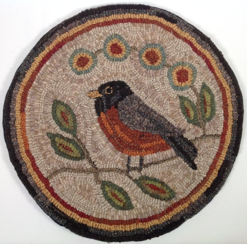 Puede incluir: Alfombra redonda, hecha a mano, con un dise&ntilde;o de p&aacute;jaro. La alfombra muestra un petirrojo con la espalda gris, el pecho naranja y la cabeza negra, posado en una rama con hojas verdes. El borde incluye tonos marrones, rojos y negros, creando una est&eacute;tica r&uacute;stica.