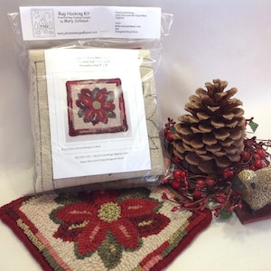 May include: A rug hooking kit with a poinsettia design, packaged in a clear bag. The kit includes a canvas and a printed pattern. A finished rug with the same design is displayed next to the kit. A large pine cone and red berries are also in the image.