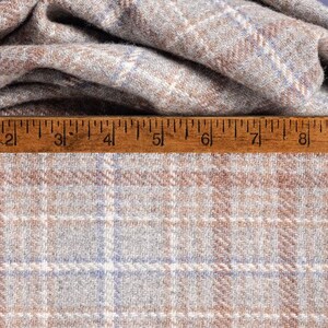 May include: Close-up of a folded fabric with a plaid pattern. The fabric is a mix of light brown, blue, and white, creating a classic plaid design. A wooden ruler is placed across the bottom of the fabric.